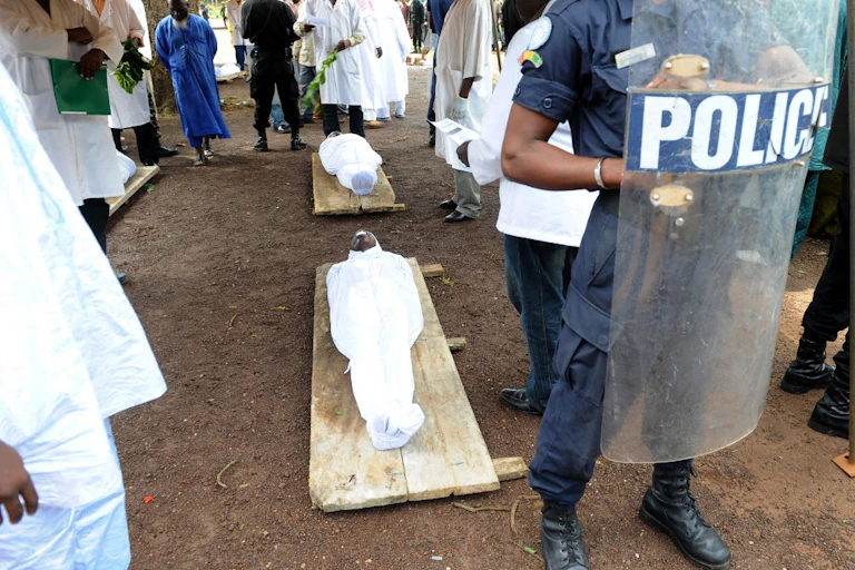Corps dans des draps blancs sur des civières derrière un bouclier antiémeute siglé police.