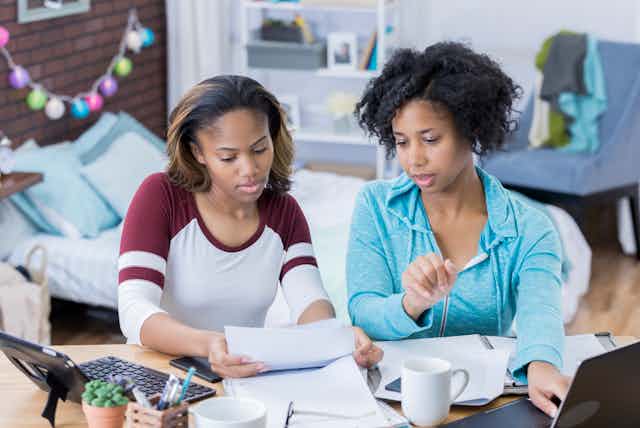 A Black mother and daughter look through paperwork while using a laptop.