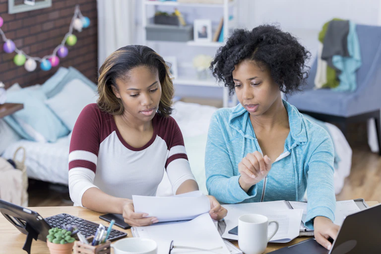 A Black mother and daughter look through paperwork while using a laptop.