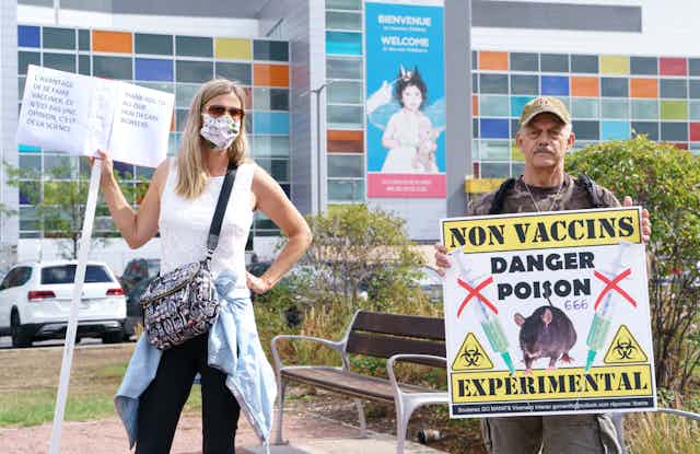 A woman stands wearing a mask and a sign that reads 'thank you to our health care workers' while a man, stands maskless with a sign that reads 'no vaccines, experimental, danger, poison, 666'