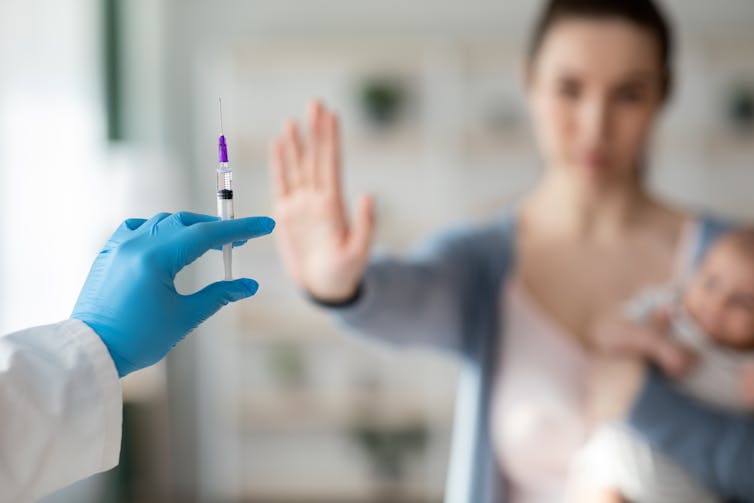 A gloved hand holds up a needle, blurred in the background a woman stands with her hand up, holding a baby