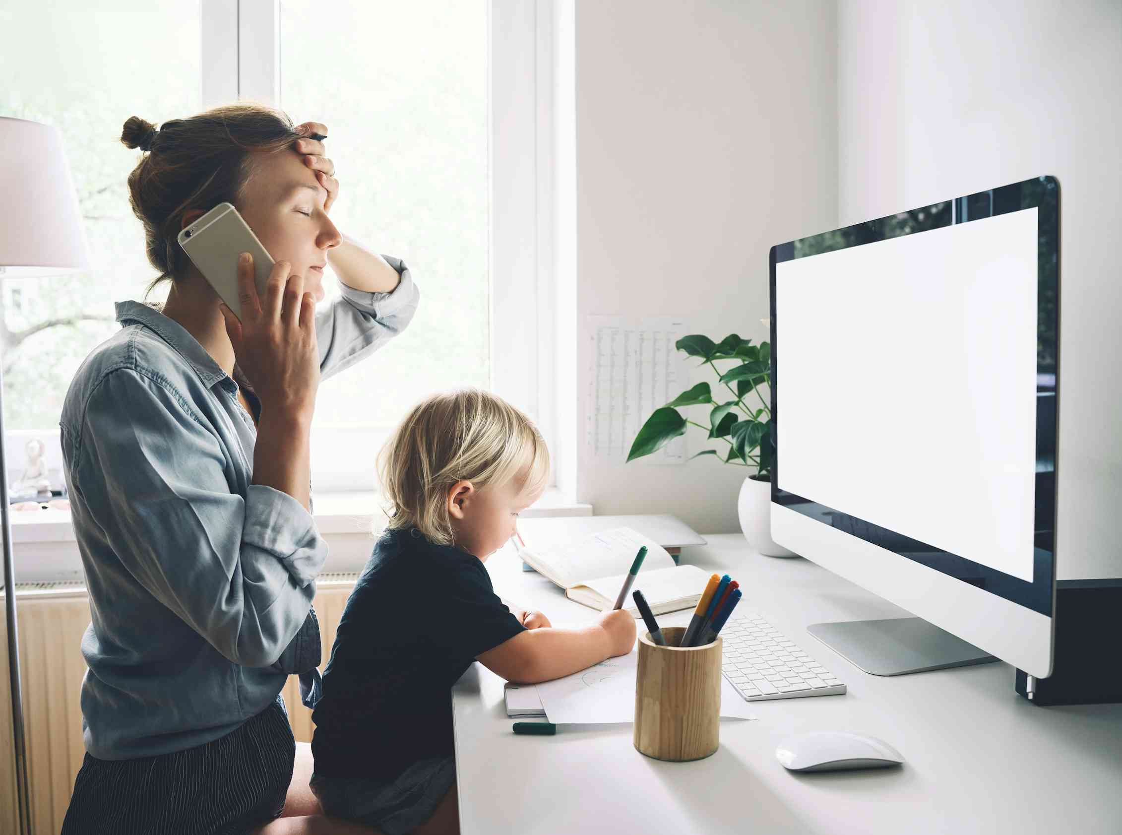 A stressed mum talks on the phone while looking at her computer, with a toddler sitting on her lap drawing