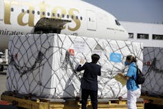 Two people inspecting a large crate covered with netting, with an airplane in the background.