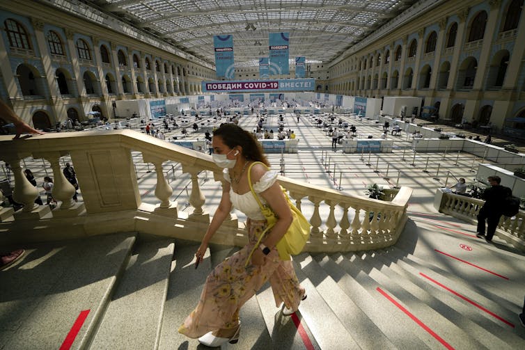 A woman climbs a curved staircase with an ornamental handrail. In the background is a vaccination clinic in a large open space with arches on the sides.