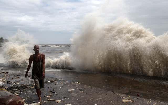 A man walks past a wave crashing on a seawall.