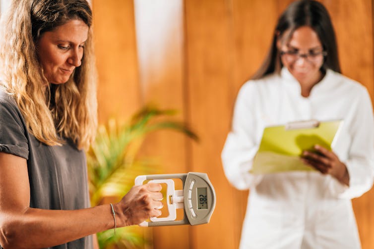 A blonde woman squeezes a device with one hand to test her hand-grip strength, while a female researcher in a white lab coat records the results.