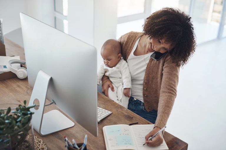 A young woman using a smartphone and computer while caring for her baby at home.