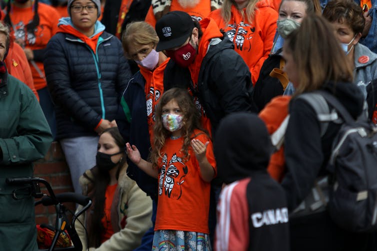 People wear orance and stand in a group, a little girl has her hands raised as a sign of respect