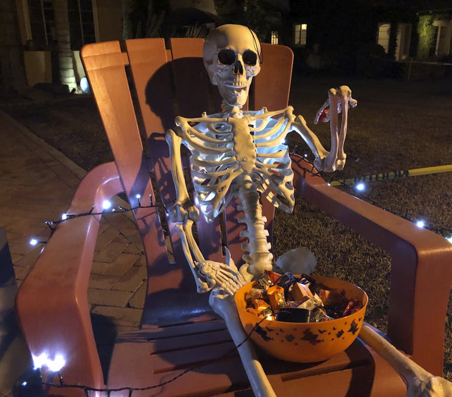 A skeleton with a candy bowl on his lap sits in an outdoor chair with lights running across his bones and a sign that reads warning electric fence
