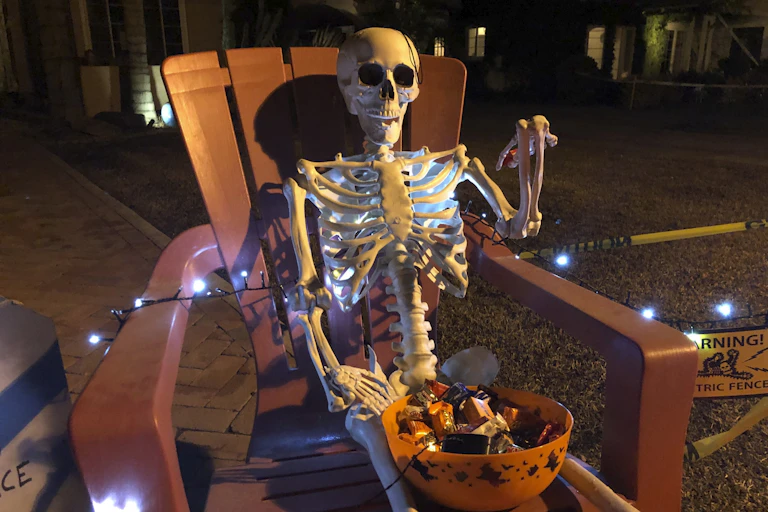 A skeleton with a candy bowl on his lap sits in an outdoor chair with lights running across his bones and a sign that reads warning electric fence