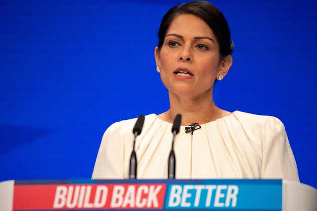 Home secretary Priti Patel speaking behind a podium at the Conservative party conference. The podium has a red and blue sign reading 'Build Back Better'