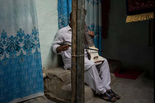 A man sits in front of a blue wall, his face is hidden behind a pillar, he poses with his dilruba