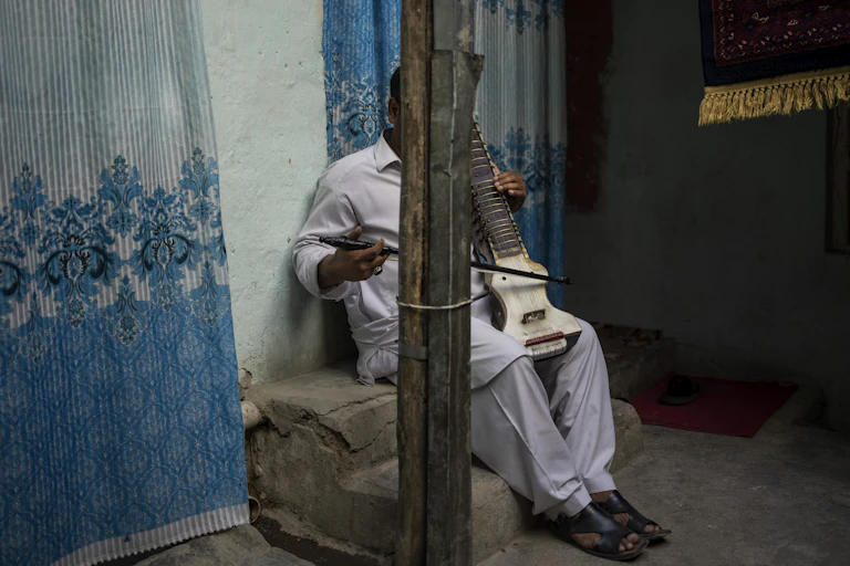 A man sits in front of a blue wall, his face is hidden behind a pillar, he poses with his dilruba