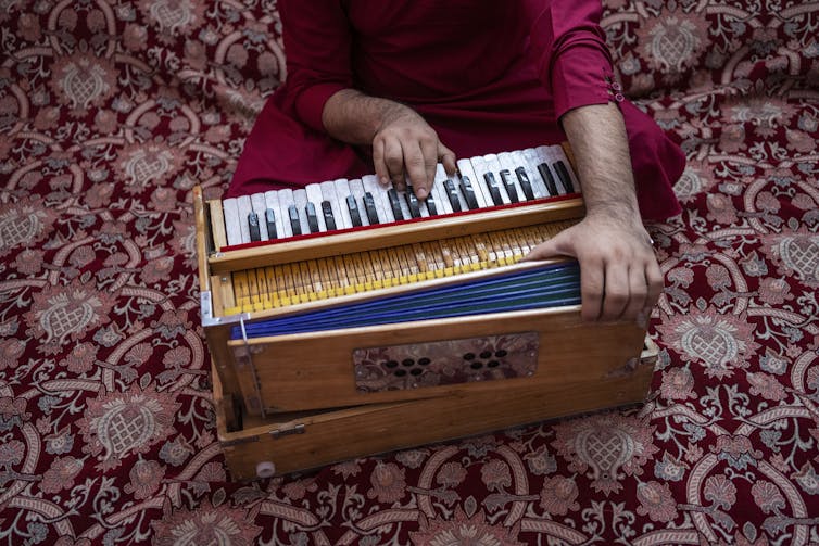 A person sits cross-legged playing the harmonium. They are wearing burgundy, sitting on a burgundy floral rug.
