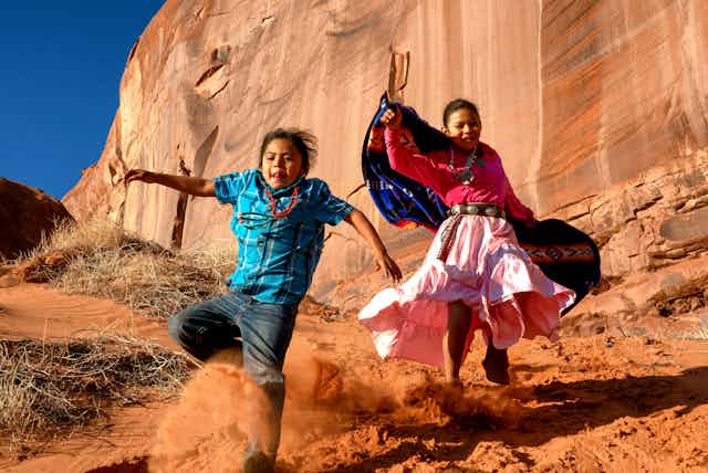 Two Native American children dressed in traditional clothing run in a desert canyon.