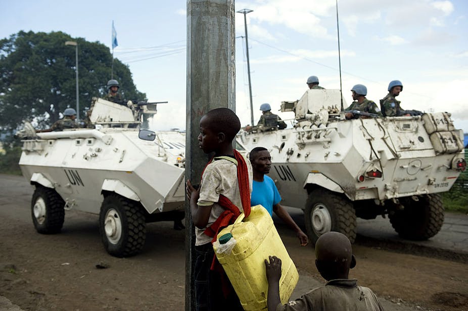 Armoured UN vehicles in the road, men wearing blue helmets on board, with some people in the foreground