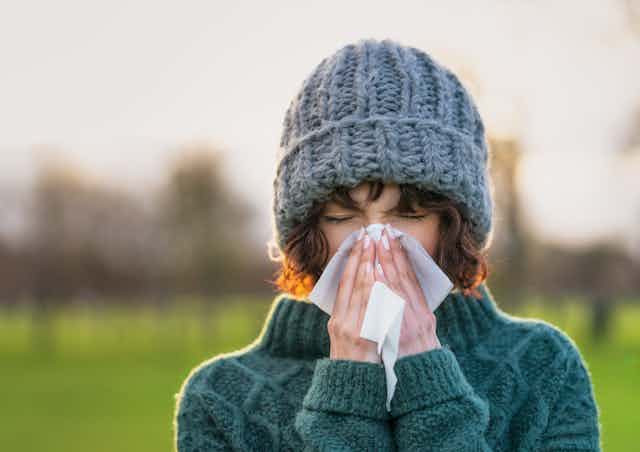 A woman, outside on a cold day, and wearing a sweater and winter cap, sneezes with a Kleenex.