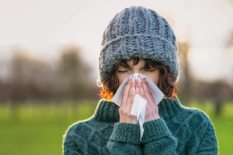 A woman, outside on a cold day, and wearing a sweater and winter cap, sneezes with a Kleenex.