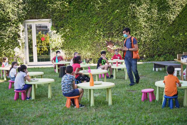 School kids in outdoor classroom with small round tables and teacher walking around.
