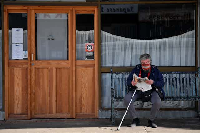 Older man in a mask and with a walking stick sits on a bench, reading the paper.