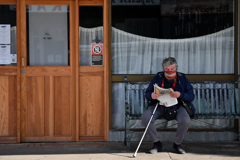 Older man in a mask and with a walking stick sits on a bench, reading the paper.
