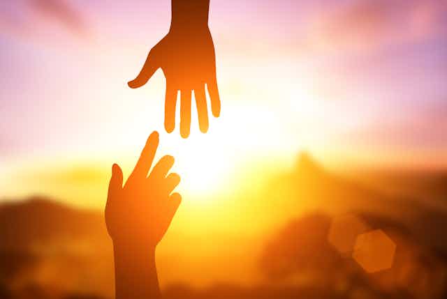 Two hands reach out to one another in silhouette with the sun shining behind them.