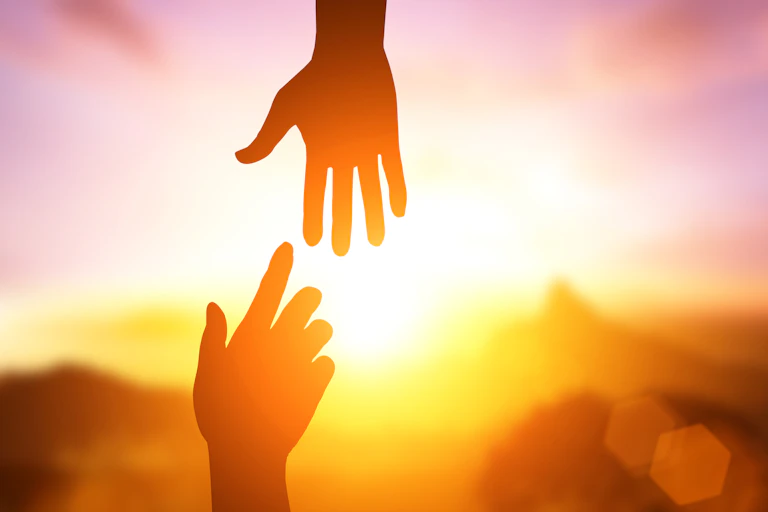 Two hands reach out to one another in silhouette with the sun shining behind them.