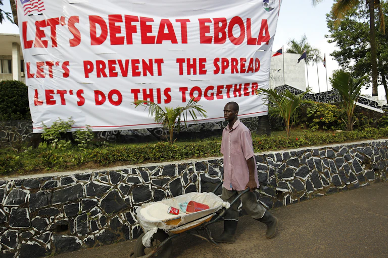 A Liberian man pushes a wheelbarrow next to a large poster with the letters: 'Let's defeat Ebola'