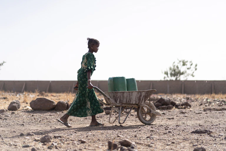 Woman pushing wheelbarrow with containers of water.