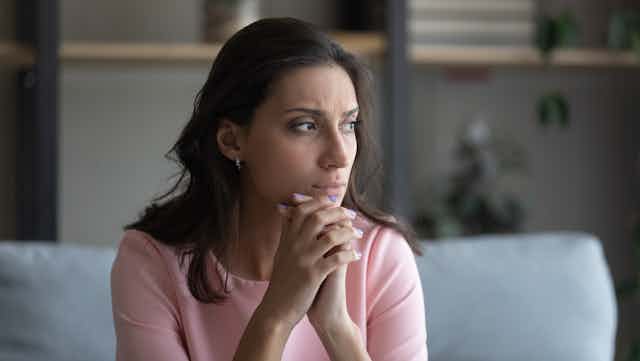 A woman sits and looks to the right with her hands clasped together below her chin.