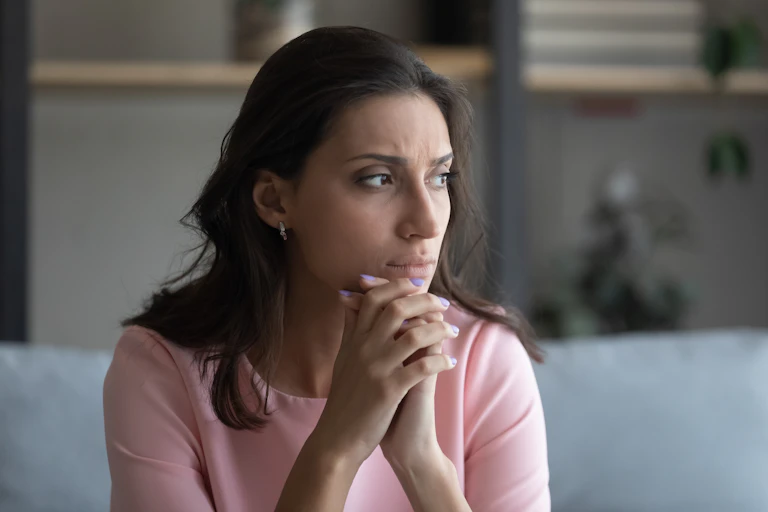 A woman sits and looks to the right with her hands clasped together below her chin.