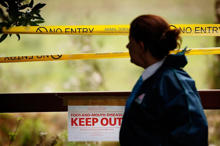 A woman stands in front of an area cordoned off with caution tape and a sign saying 'Keep Out.'