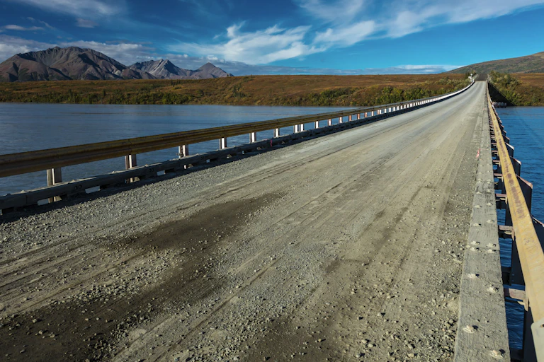 A long, gravel-covered bridge with low side rails and mountains in the distance