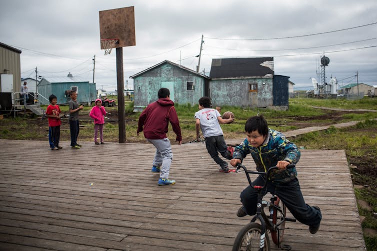 Children play basketball and ride bikes on a large wooden platform over a wet field.  The weather-beaten houses stand in the background.