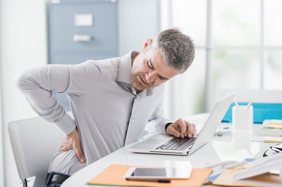 A man sitting at his desk working on his laptop presses a hand onto his sore lower back in pain.