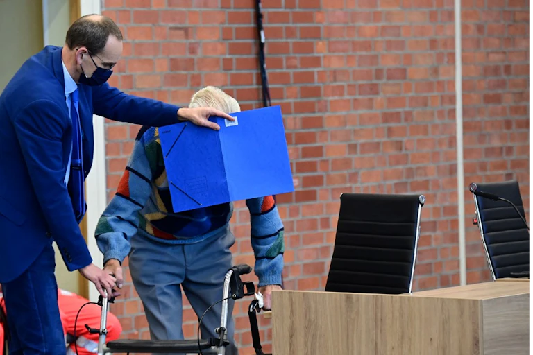 Un viel homme se tenant à son déambulateur quitte une salle d'audience le visage caché par son avocat au moyen d'une pochette en carton bleu.