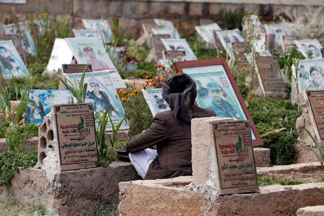A Houthi rebel visits the grave of a relative killed in the civil war in Yemen. In the background are graves, many of which have photographs of the people buried there.