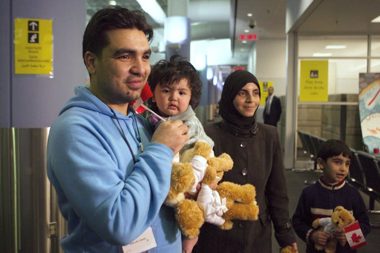 A man holds a baby and two teddy bears and smiles while his smiling wife and son, holding a small Canadian flag, stand beside him.