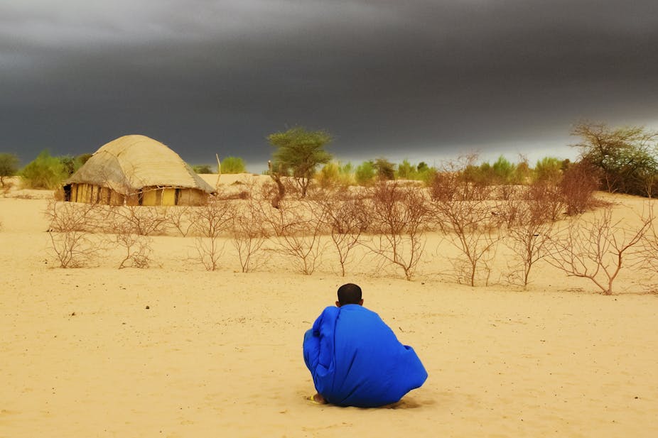 A man in blue overall sitting in the desert in Timbuktu, Mali, looking at dark clouds waiting for the rain.
