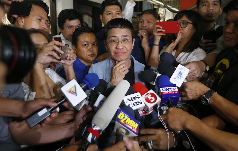 Maria Ressa smiles at the camera while surrounded by TV news reporters and microphones.