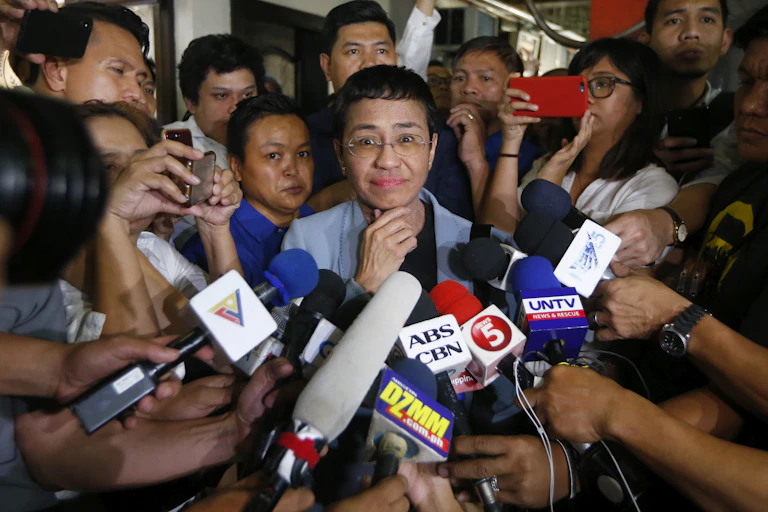 Maria Ressa smiles at the camera while surrounded by TV news reporters and microphones.