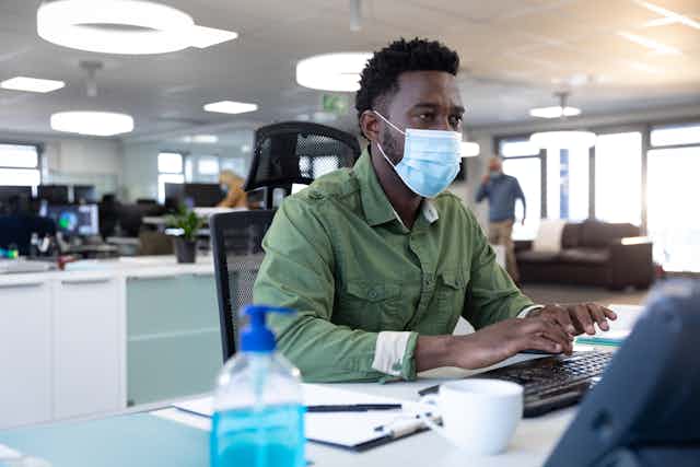 African-Australian man in his thirties wearing a mask types at a computer in a busy office.