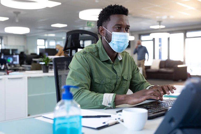 African-Australian man in his thirties wearing a mask types at a computer in a busy office.