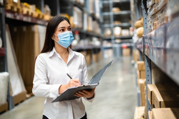 Masked woman with a clipboard surveys a storeroom.