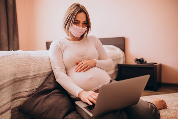 Pregnant woman in mask sits on bedroom floor, looking at laptop.