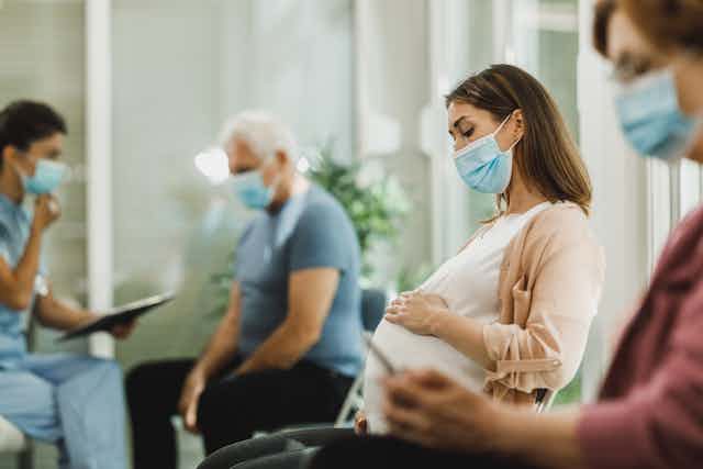 Pregnant woman sits in a medical waiting room, surrounded by masked patients.
