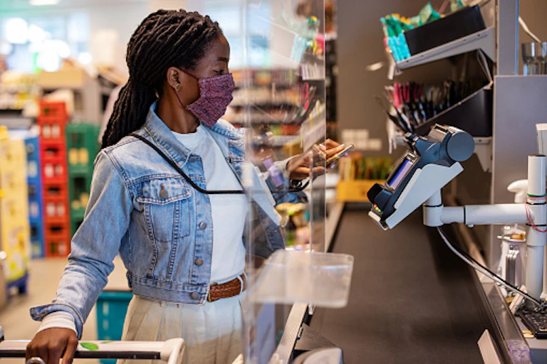 A woman wearing nosemask and holding a mobile phone over a POS machine