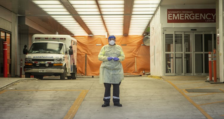 A paramedic in personal protective equipment stands in an ambulance bay with an ambulance on the left and the entrance to Emergency on the right