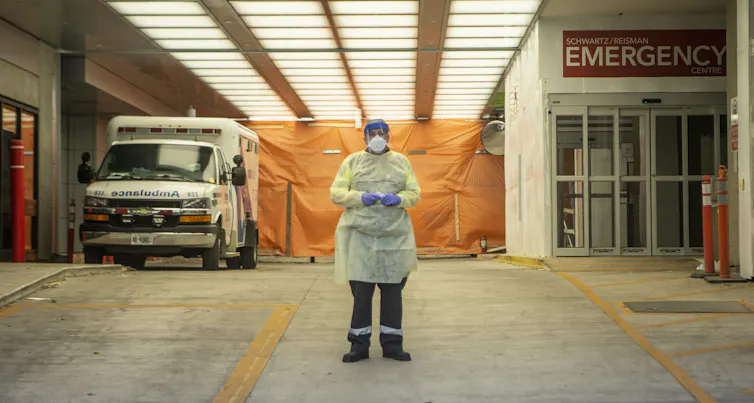 A paramedic in personal protective equipment stands in an ambulance bay with an ambulance on the left and the entrance to Emergency on the right