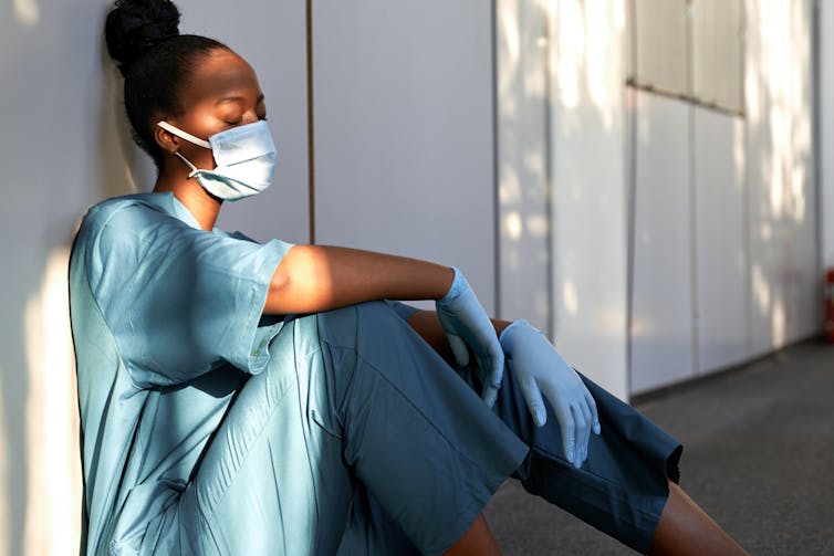 A health-care worker wearing scrubs, gloves and a face mask sits on the floor leaning against a wall